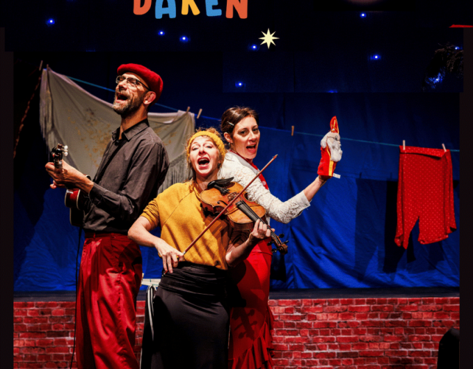 Drie muzikanten staan op een podium en zingen terwijl ze instrumenten bespelen. De achtergrond toont een nachtelijke scène met sterrenlichtjes, een ronde lamp als maan en wasgoed aan een lijn. Bovenaan staat de titel “Luister naar de daken”.